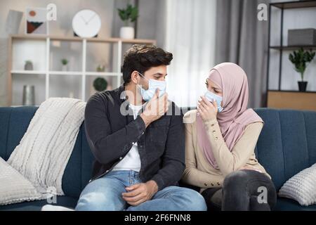 Nahaufnahme eines jungen, angenehmen arabischen Paares in Gesichtsschutzmasken, das zu Hause im gemütlichen Wohnzimmer zusammensitzt und sich gegenseitig anschaut. Selbstisolationskonzept, Quarantäne, covid-19. Stockfoto
