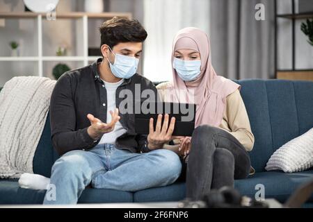 Hübscher arabischer Mann in Schutzmaske, redet und gestikuliert mit seiner muslimischen Freundin in Hijab und Maske, sitzt zu Hause und diskutiert Informationen, die sie von der Tablette lesen. Covid-19 Isolationskonzept. Stockfoto