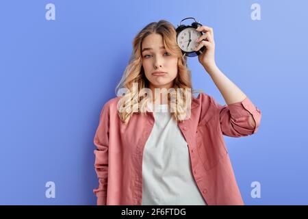 Verschlafene Frau mit Wecker am Kopf befestigt, junge Studentin in Freizeitkleidung mit frustrierten Gesicht Blick auf die Kamera, wollen nicht zur Universität gehen. Isolieren Stockfoto