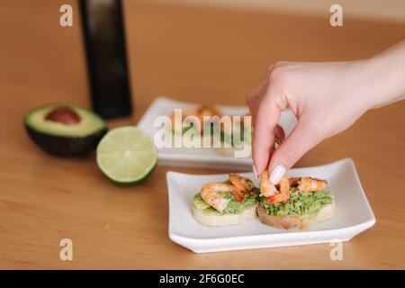 Die weibliche Hand gab Garnelen auf Bruschetta Toast mit Avocado. Gesunde Vorspeise aus Bruschetta mit Guacamole-Garnelen Stockfoto