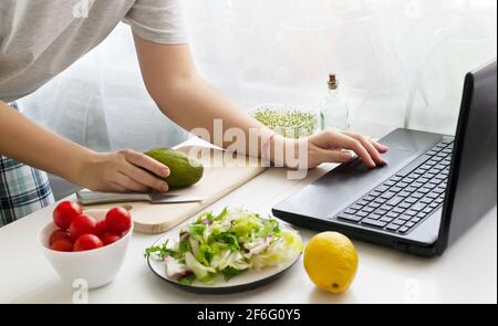 Eine Frau hält eine Avocado in der Hand und sucht auf ihrem Laptop nach einem Rezept für einen veganen Salat. Salat und Zutaten stehen auf dem Tisch. Online-Kochen Stockfoto