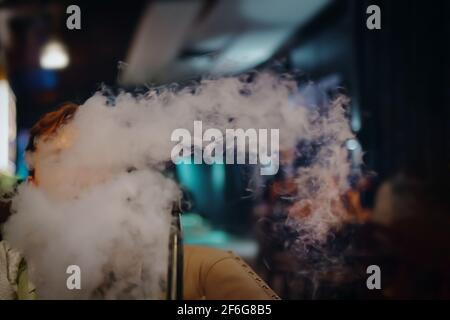 Junger Mann im Café raucht Shisha. Dicker Rauch von Shisha. Mann auf dem Sofa im Restaurant. Neonschilder auf dem Hintergrund. Entspannen Sie sich in der Shisha Bar. Stockfoto