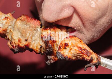 Ein Mann isst ein saftig duftendes Stück Schweinekebab auf einem Spieß. Fetthaltige, kalorienreiche Lebensmittel. Nahaufnahme. Stockfoto