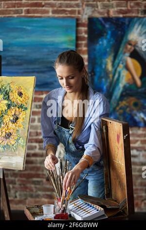 Maler Auswahl Kunst Pinsel für die Arbeit. Porträt einer jungen lächelnden Künstlerin im Kunststudio. Kreativer Prozess, Entspannung, Freizeit, Hobby, Stress Stockfoto
