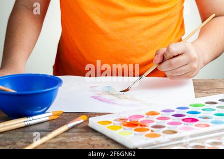 Ein kaukasischer Junge zeichnet ein abstraktes Bild auf einem weißen Papier mit einem Satz Aquarell. Er ist Linkshänder und hält Pinsel. Künstlerische Entwicklung, Stockfoto