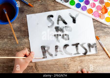 Das kaukasische Kind schreibt mit Pinsel und schwarzem Aquarell auf einem weißen Papier „Say No to Racism“-Slogan. Ein Kind, das sich sozialer Angelegenheiten bewusst ist Stockfoto
