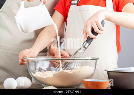 Mutter und Tochter kochen gemeinsam im Küchenkonzept mit Das Mädchen hält einen Schneebesen und die Frau gießt vorsichtig Milch aus dem Krug in den m Stockfoto