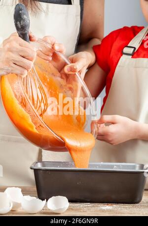 Mutter und kleine Tochter kochen zusammen Konzept mit einer Glasschüssel Orangenkuchen Mischung gegossen in eine Laib Pfanne zu backen. Beide tragen Apro Stockfoto