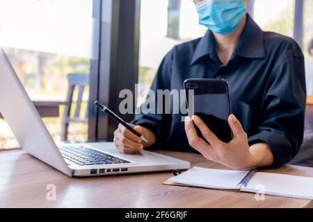 Junge Frau mit schützender Gesichtsmaske sitzt im Café am Holztisch mit Smartphone.und Laptop. Mädchen im Internet surfen, chatten, bloggen. Fe Stockfoto
