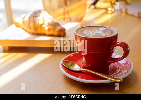 Nahaufnahme von heißem Cappuccino, Latte, Caramel Macchiato auf dem Tisch mit Blur Coffee Shop Hintergrund. Heißer Cappuccino-Kaffee auf Holztisch mit Blur Croissant b Stockfoto