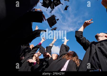 Gratulation aus der Perspektive einer glücklichen Gruppe von sechs jungen, fröhlichen Absolventen in schwarzen Kleidern, die ihre Kopfbedeckung in die Luft werfen und feiern, Stockfoto