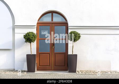 Geschlossene rote Holztür mit zwei dekorativen Bäumen in Töpfen an einer weißen Wand der Gebäudefassade. Restaurant, vorübergehend während Coronavirus pandemi geschlossen Stockfoto