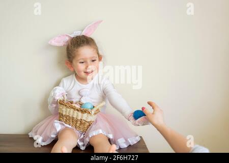 Selektiver Fokus. Kleines süßes Mädchen im Osterhasen-Kostüm sitzt lächelnd vor der Wand. Feiern mit einem Korb von Ostern e Stockfoto