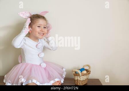 Kleines süßes Mädchen im Osterhasen-Kostüm sitzt lächelnd vor der Wand. Feiern mit einem Korb mit Ostereiern in der Hand. Auswahl Stockfoto