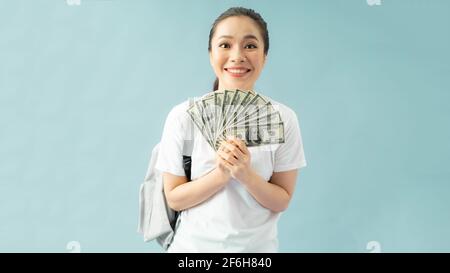 Schockiert Mädchen Studentin im Rucksack isoliert auf pastellblauem Hintergrund, halten Fan von Bargeld in Dollar-Banknoten Hand auf Wange Stockfoto