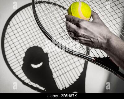 Eine Person mit einem Tennisball in der Hand und Ein Tennisschläger wirft einen Schatten auf eine sonnendurchflutete Oberfläche Stockfoto
