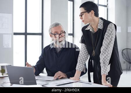Reifer selbstbewusster Geschäftsmann sitzt am Tisch mit Laptop und Im Gespräch mit seinem Partner während des Arbeitstages im Büro Stockfoto