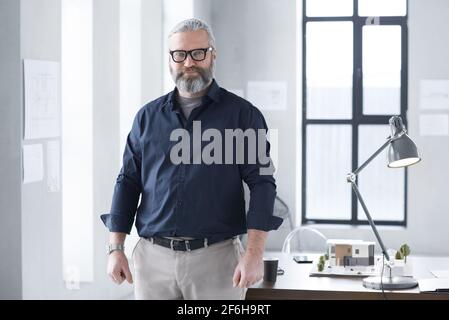 Porträt eines reifen bärtigen Architekten, der während der Arbeit die Kamera anschaut Im Büro mit Architekturmodell Stockfoto