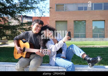 Ein junges lateinisches Paar, das auf dem Universitätscampus ein Selfie macht, singt und Gitarre spielt. Universitätsleben, tausendjährige Generation. Stockfoto