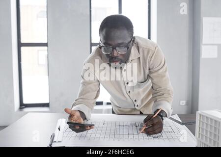 Afrikanischer junger Architekt, der sein Mobiltelefon verwendet, während er Blaupause zeichnet Am Tisch im Büro Stockfoto