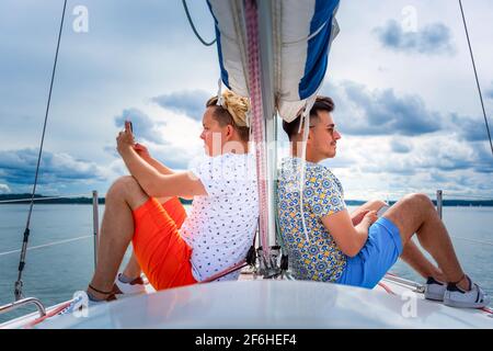 Zwei junge Mann sitzen auf einem Segelboot Deck Bogen adn Blick auf gegenüberliegenden Seiten. homosexuell paar Urlaub auf einer Yacht Stockfoto