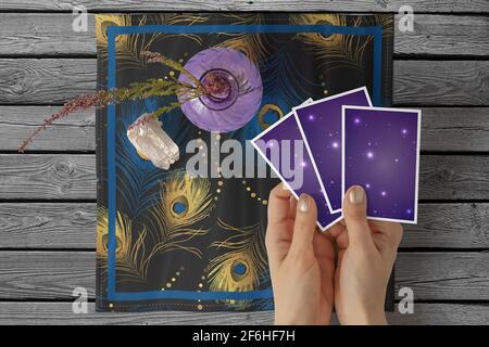 Hände einer jungen Frau mit goldenem Nagellack, die drei Tarot- oder orakelkarten auf einem quadratischen Tischtuch mit schwarzem Pfauenfedermuster hält Stockfoto