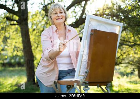 Portrait einer positiv talentierten Künstlerin mittleren Alters, die im grünen Park vor der Staffelei steht, mit Pinsel hält und an einem Bild arbeitet. Kreativität, Inspiration, Kunst und Malkonzept Stockfoto