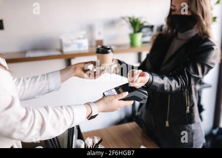 Frau mit Gesichtsmaske, die im Café serviert und nach dem Lockdown geöffnet wurde. Frau zahlt mit Smartphone und nfc-Technologie. Nahaufnahme der menschlichen Hände, Herstellung mit Hilfe von Mitteln Stockfoto