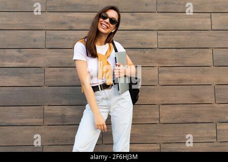 Faszinierende schöne lächelnde charmante junge Brunet Mädchen Blick auf die Kamera Halten Computer Laptop und schwarze Sonnenbrille in weißem T-Shirt und Leicht Stockfoto