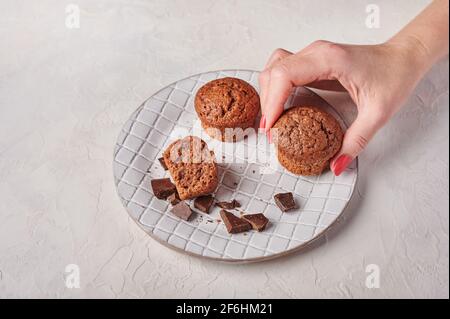 Die Hand der Frau hält einen Cupcake, andere und Schokoladenstücke in strukturierter Platte auf hellem Holzhintergrund, selektiver Fokus Stockfoto