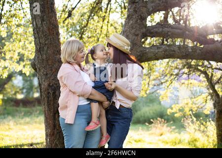 Familie mit mehreren Generationen im Garten im Sommer. Die Großmutter im Ruhestand hält ein schönes kleines Mädchen an den Händen, während sie mit der jungen hübschen Mutter flattert und die Nase berührt. Stockfoto