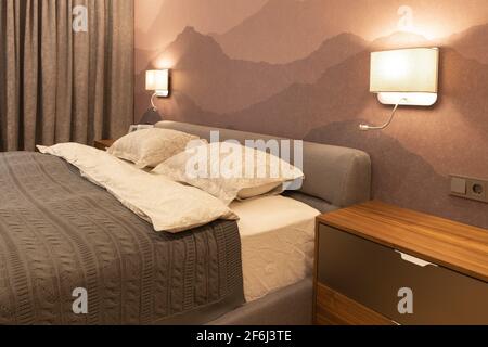 Modernes Schlafzimmer in der Wohnung, in neutralen Erdtönen. Doppelbett mit einem weichen textilen Kopfteil. Schöne Nachttischlampen leuchten mit warmem Licht. Min Stockfoto