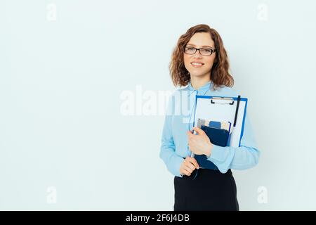 Selbstbewusst lächelnde junge Geschäftsfrau mit Brille, blauem Hemd und schwarzem Rock mit Klemmbrett, Papieren und Notizblock auf weißem Studiorücken Stockfoto