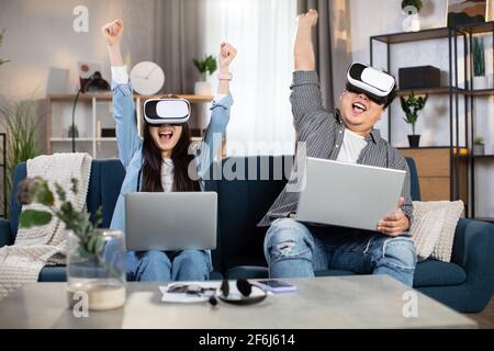 Fröhliche asiatische Familie sitzt zusammen auf dem Sofa mit erhobenen Armen und mit einem modernen Laptop mit VR-Brille. Junges Paar, das Zeit zu Hause mit Spaß und Vergnügen verbringt. Stockfoto