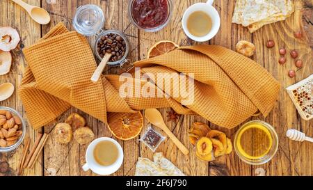 Zusammensetzung der Frühstückssnacks mit einem gefalteten Küchentuch, um das Pfannkuchen, Obstchips, Nüsse, Honig und zwei Tassen Kaffee herum gelegt werden. Gesunder Snack und Öko f Stockfoto