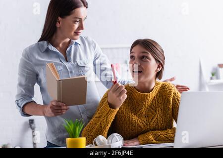 Teenager-Mädchen denken und halten Stift, während Hausaufgaben in der Nähe Mutter mit Buch Stockfoto