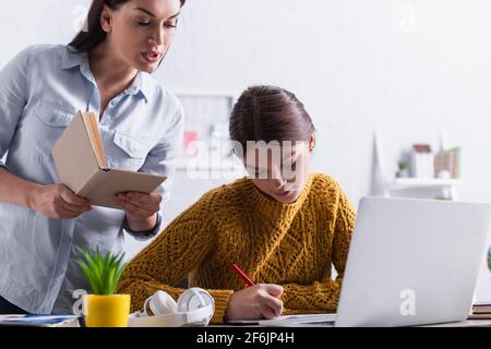 Teenager-Mädchen schreiben, während Hausaufgaben in der Nähe Mutter mit Buch Und Laptop Stockfoto