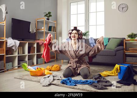 Müde Hausfrau, die Kleidung vom Boden abholt und sich darüber beschwert Ständiges Chaos zu Hause Stockfoto