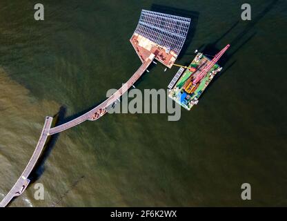 22. März 2021, Mecklenburg-Vorpommern, Koserow: Neben der im Bau befindlichen neuen Seebrücke auf der Insel Usedom liegt ein schwimmender Ponton mit Kran. Durch den notwendigen Austausch von als Laufplanken verlegtem Tropenholz und die erneute Verzinkung von Geländersegmenten wird die Brücke nun erst Anfang Juli 2021 für das Brückenfest bereit sein. Der Pier erstreckt sich fast 300 Meter in die Ostsee, und die geschwungene Wellenform des geraden Piers auf anderen Brücken ist auffällig. Der Neubau wird mehr als fünf Millionen Euro kosten. (Foto aufgenommen mit einer Drohne) Foto: Jens Büt Stockfoto