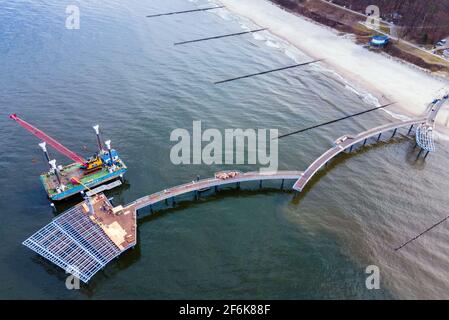 22. März 2021, Mecklenburg-Vorpommern, Koserow: Neben der im Bau befindlichen neuen Seebrücke auf der Insel Usedom liegt ein schwimmender Ponton mit Kran. Durch den notwendigen Austausch von als Laufplanken verlegtem Tropenholz und die erneute Verzinkung von Geländersegmenten wird die Brücke nun erst Anfang Juli 2021 für das Brückenfest bereit sein. Der Pier erstreckt sich fast 300 Meter in die Ostsee, und die geschwungene Wellenform des geraden Piers auf anderen Brücken ist auffällig. Der Neubau wird mehr als fünf Millionen Euro kosten. (Foto aufgenommen mit einer Drohne) Foto: Jens Büt Stockfoto