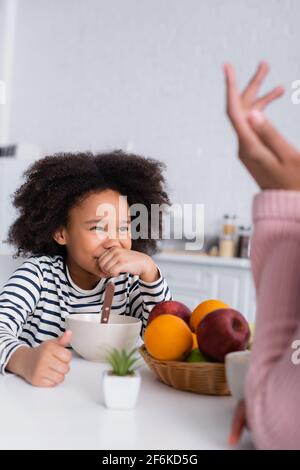 Lachendes afroamerikanisches Mädchen, das den Mund bedeckt, während es die Mutter anschaut Gestikulieren auf verschwommenem Vordergrund Stockfoto