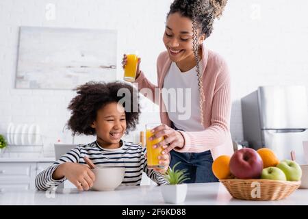 Lächelnde afroamerikanische Frau, die der Tochter in der Nähe Orangensaft gab Früchte auf verschwommenem Vordergrund Stockfoto