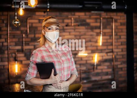 Bestellung in einem Restaurant. In einem Restaurant und steht ein Porträt einer in Pastellfarben gekleideten Kellnerin und Gastgeberin mit einer schützenden Gesichtsmaske Stockfoto