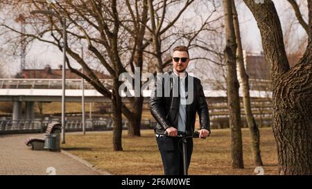 Der junge Mann in einer schwarzen Jacke und Sonnenbrille reitet eine Elektronischer Roller Stockfoto