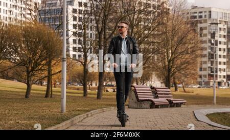 Der junge Mann in einer schwarzen Jacke und Sonnenbrille reitet eine Elektronischer Roller Stockfoto