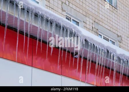 Viele Eiszapfen hängen vom Dach. Im Hintergrund ist eine Backsteinmauer eines Wohnhauses mit Fenstern. Stockfoto