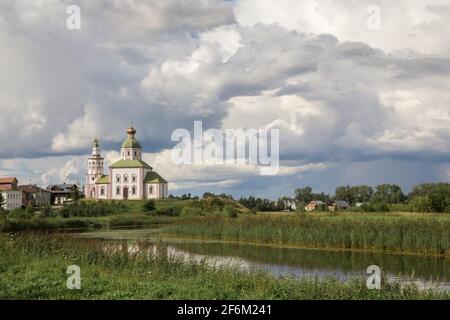 Eine alte Kirche aus weißem Stein am Ufer eines kleinen Flusses. Es gibt einstöckige Wohngebäude in der Nähe des Tempels. Von oben hängen dicke Wolken. Stockfoto