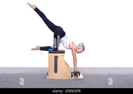 Eine 20-jährige Trainerin übt Pilates auf einem Fahrstuhlstuhl und hebt ihr Bein hoch über ihren Kopf. Eine Frau ruht auf ihrem Knie und ihren Händen Stockfoto