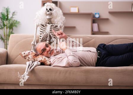 Alter Mann sitzt auf dem Sofa mit dem weiblichen Skelett In unsterbst Liebe Konzept Stockfoto