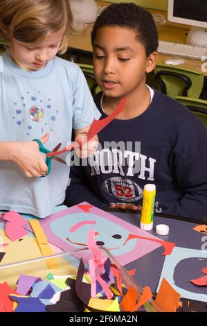 Unabhängige Grundschule Klasse 4 und Kindergarten Buddies Mädchen mit Schere, um Papier für die Collage zu schneiden, wie die ältere Studente sie unterstützt Kunstaktivität Stockfoto